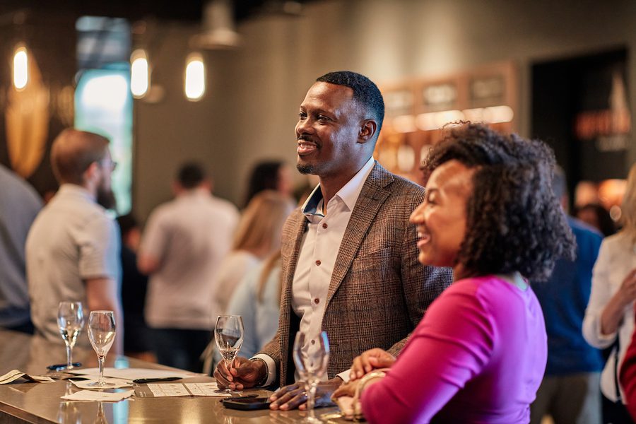 Smiling people chatting at a lively indoor reception, with a man in a blazer and a woman in a pink top standing at a table with wine glasses and menus