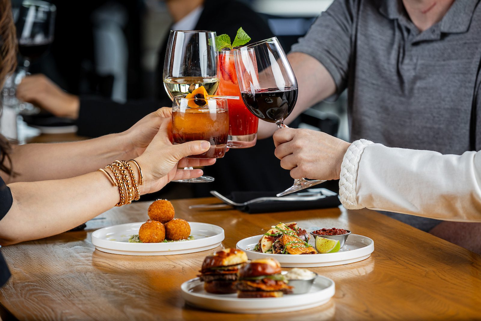 Hands toasting with wine and colorful cocktails over small plates of tapas and appetizers at a restaurant table