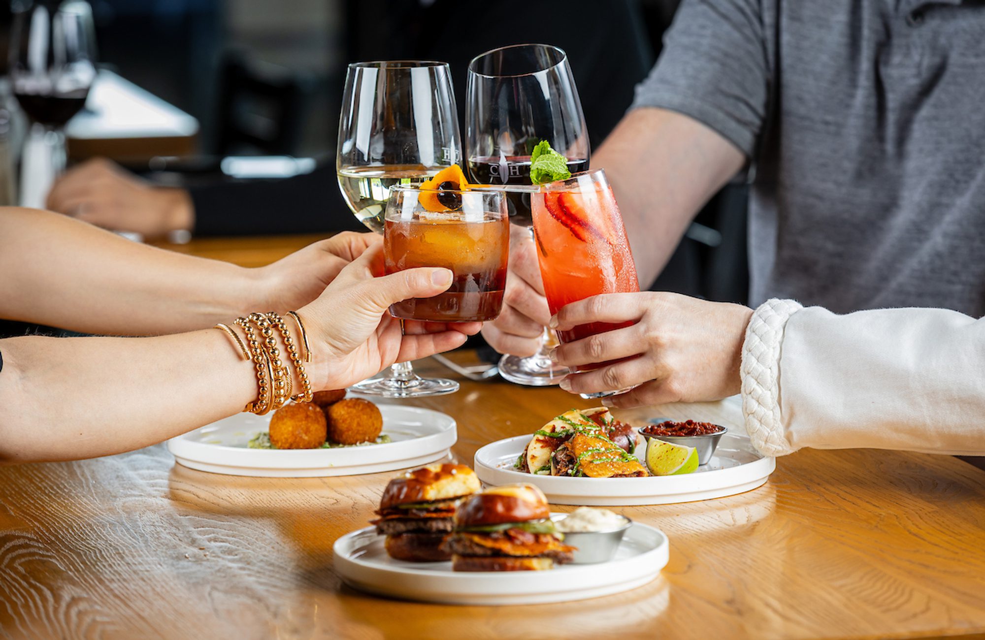 Hands clink cocktails and wine glasses over a wooden table with shared plates of appetizers and small bites in a restaurant