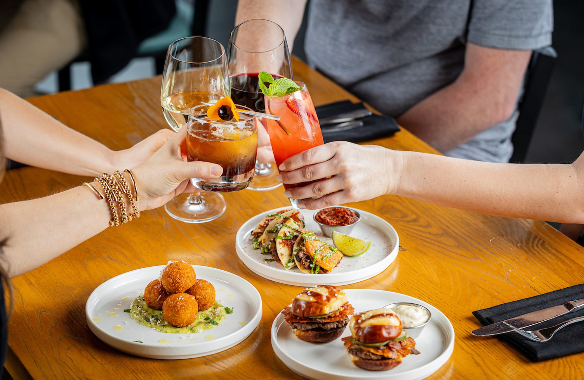 People clinking cocktails over a table with small plates of appetizers: mini tacos, fried croquettes, and sliders.