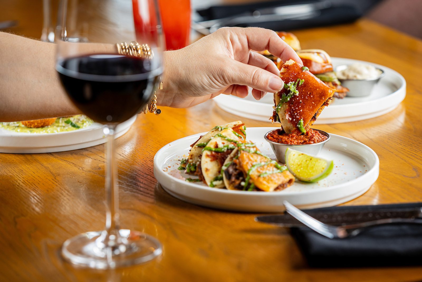 Hand reaching to dip a crispy taco into red salsa on a white plate, with lime wedge and a wine glass in the foreground at a restaurant table