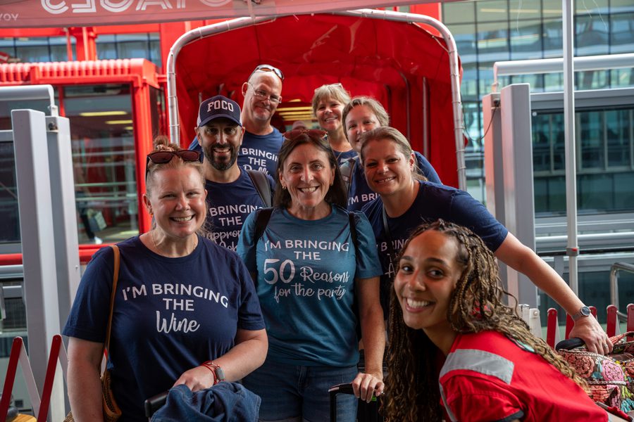 Smiling group of seven travelers posing with luggage in a red shuttle, wearing matching “I’m bringing the wine” party shirts outside a modern terminal