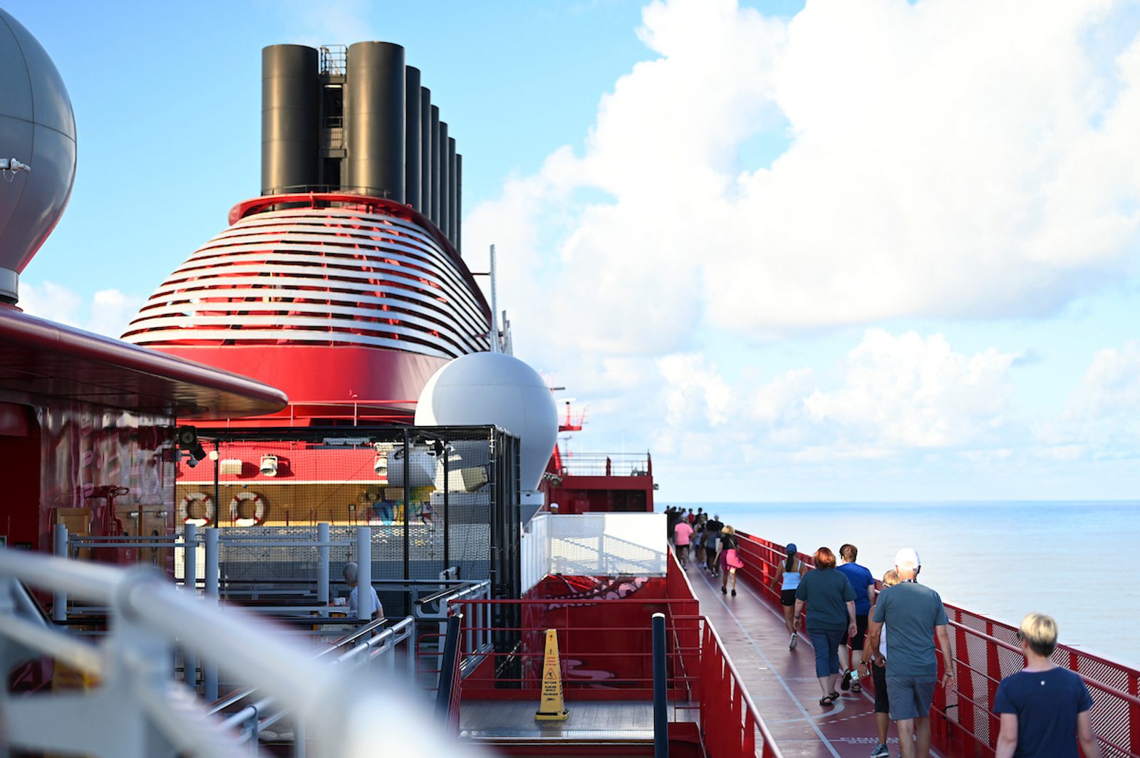 Passengers walk along a red cruise ship deck beside the ocean, with a large red-and-white funnel and bright cloudy sky overhead