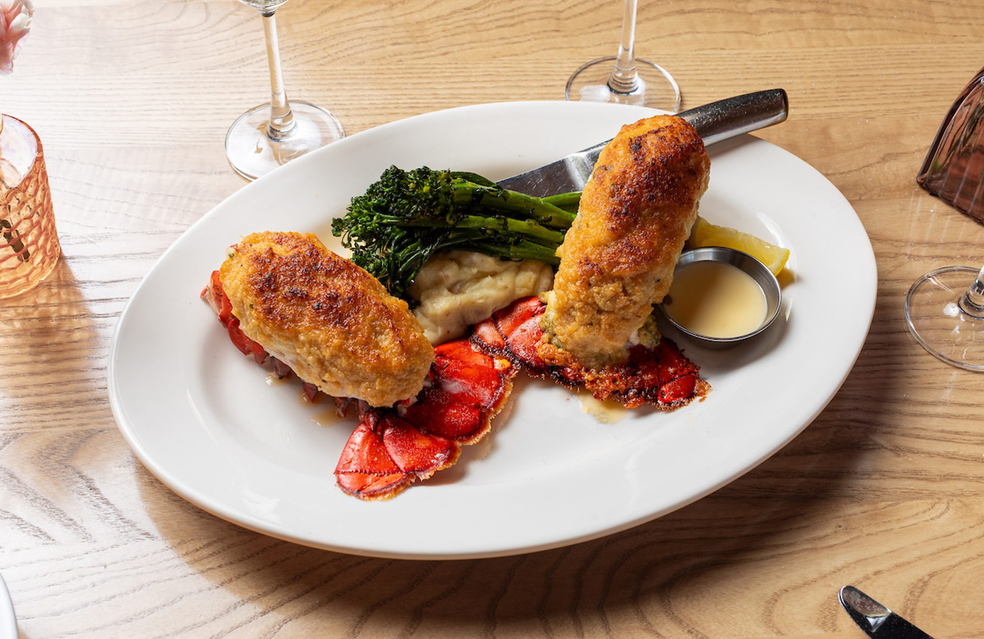 Two stuffed lobster tails on lobster shells with mashed potatoes, broccolini, lemon wedge and dipping sauce on plate.