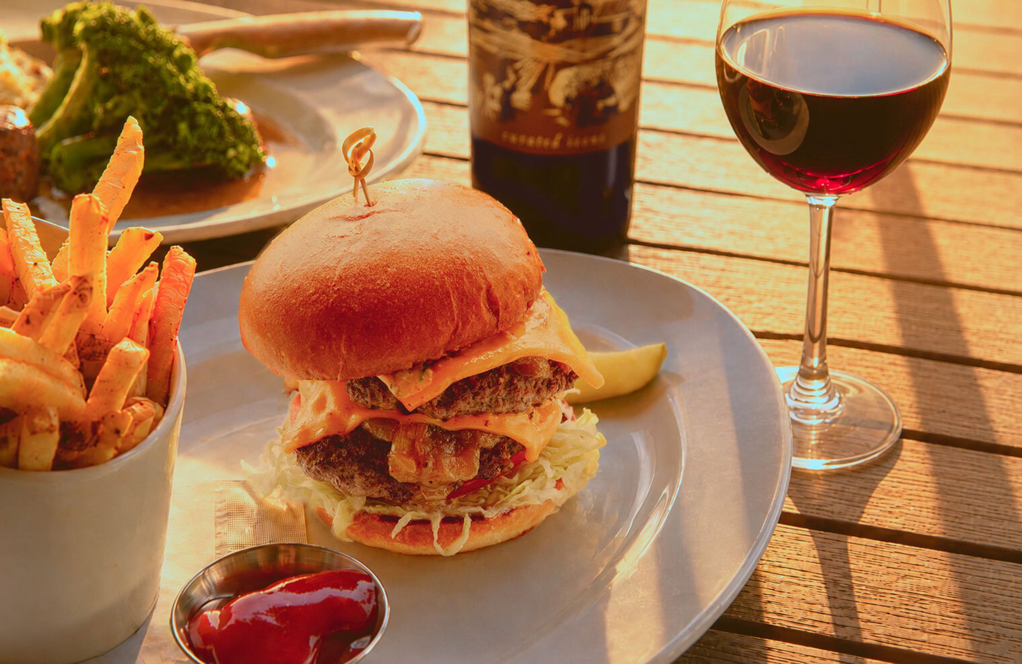 Double cheeseburger with shredded lettuce and pickle on plate, cup of fries with ketchup, glass of red wine on wooden table.