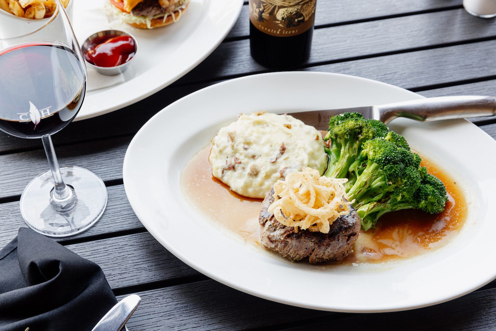 Steak with fried onion strings, mashed potatoes and broccoli in brown sauce on a white plate; glass of red wine nearby.