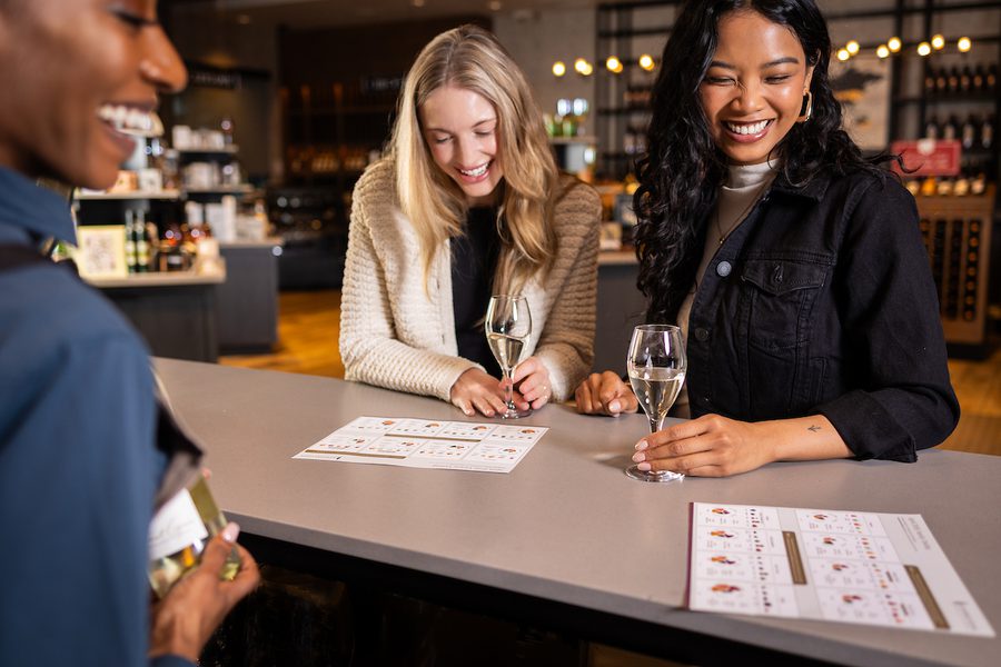 Three people smiling at a wine bar, holding champagne flutes and looking over tasting menus on the counter in a warmly lit shop
