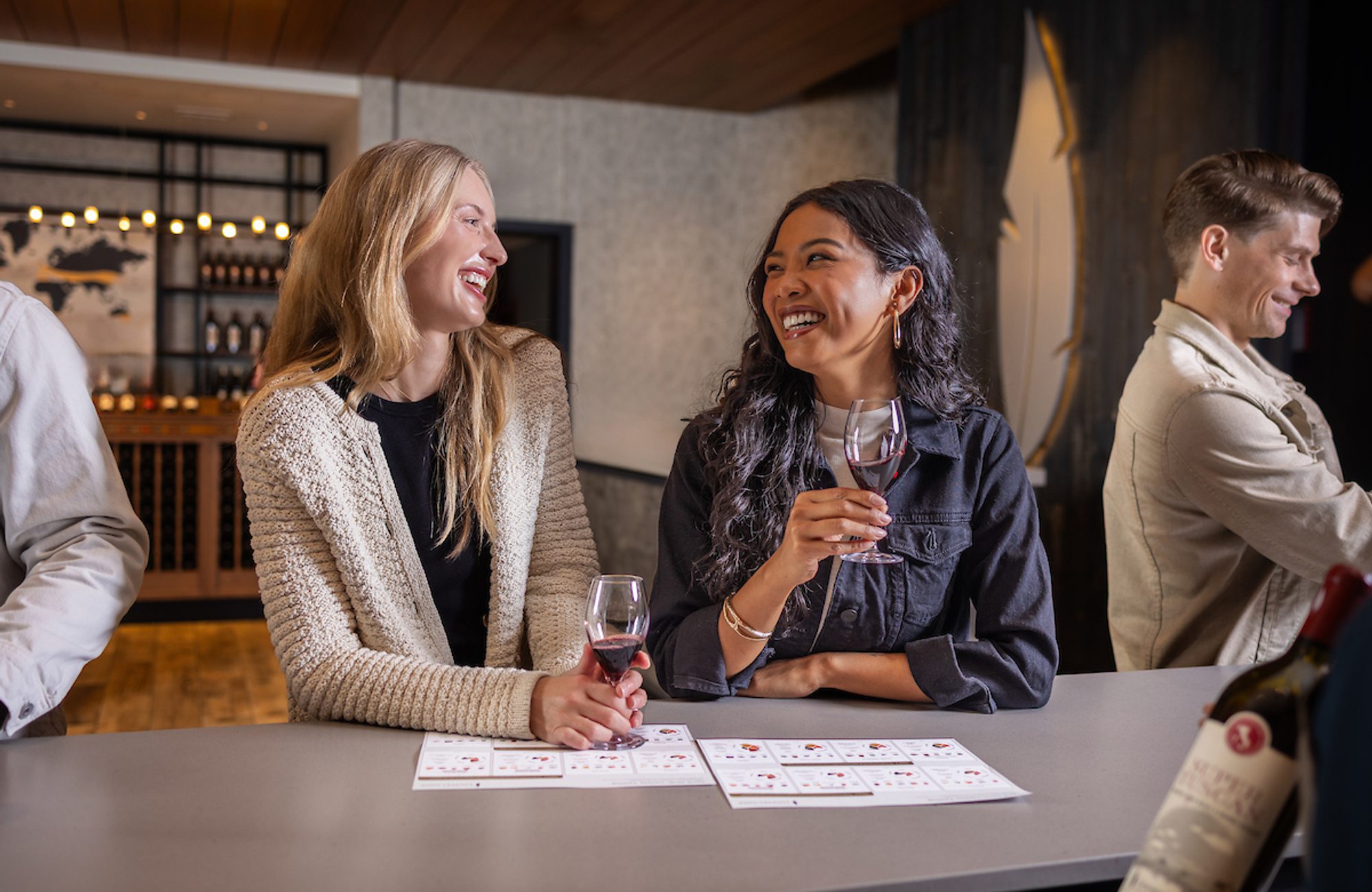 Two people smiling and chatting at a winery bar, each holding a glass of red wine, with tasting menus on the counter and others in the background