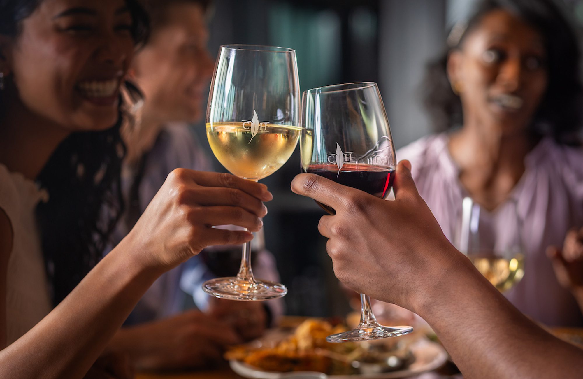 People clink wine glasses in a lively dinner toast, with plates of food and smiling friends softly blurred in the background.