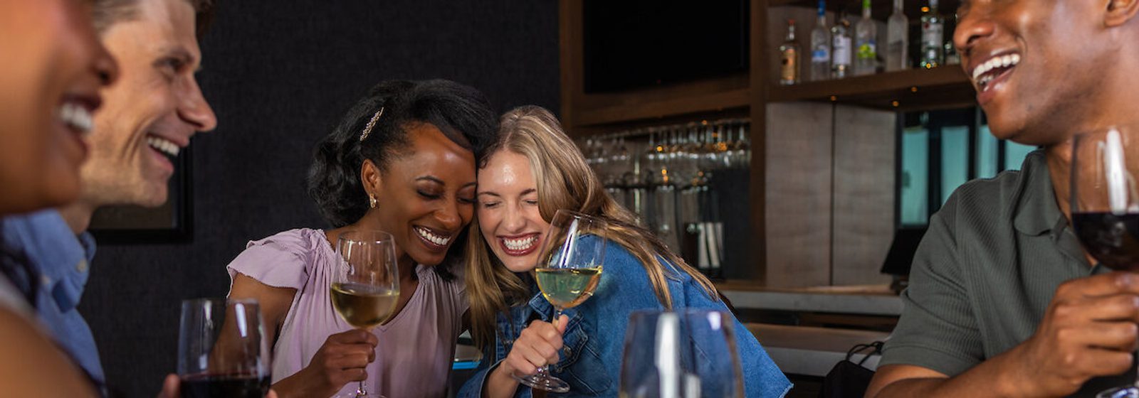 Friends laughing and sharing wine glasses at a bar, with two women at the center hugging and smiling while others chat in the foreground and background