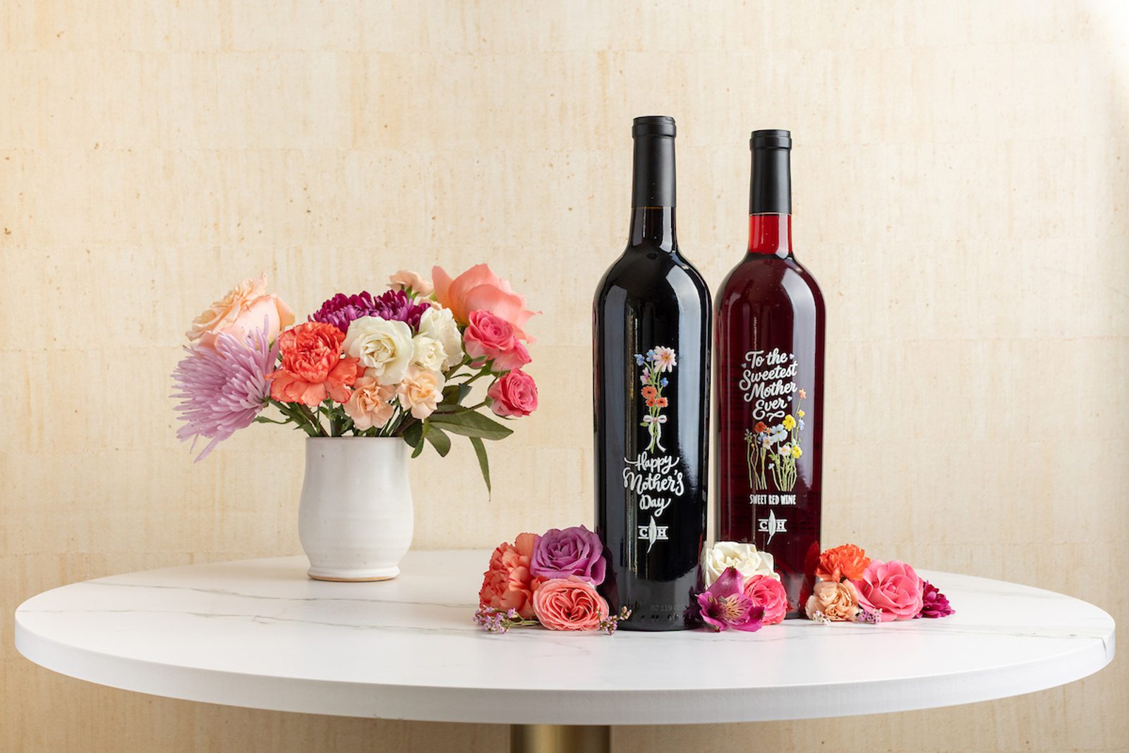 Two wine bottles with labels reading Happy Mother's Day and To the Sweetest Mother Ever, next to a vase of flowers.