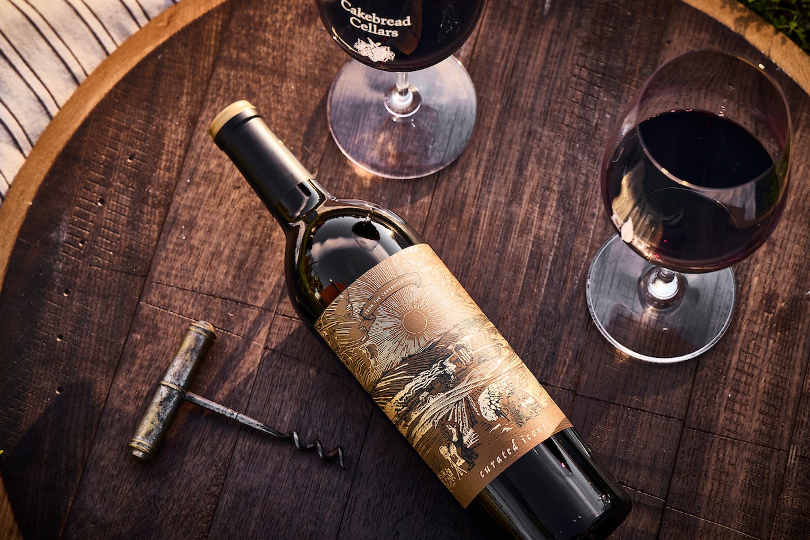 Red wine bottle with gold illustrated label reading "curated icons", two filled glasses (one labeled "Cakebread Cellars") and a corkscrew on a wooden barrel.