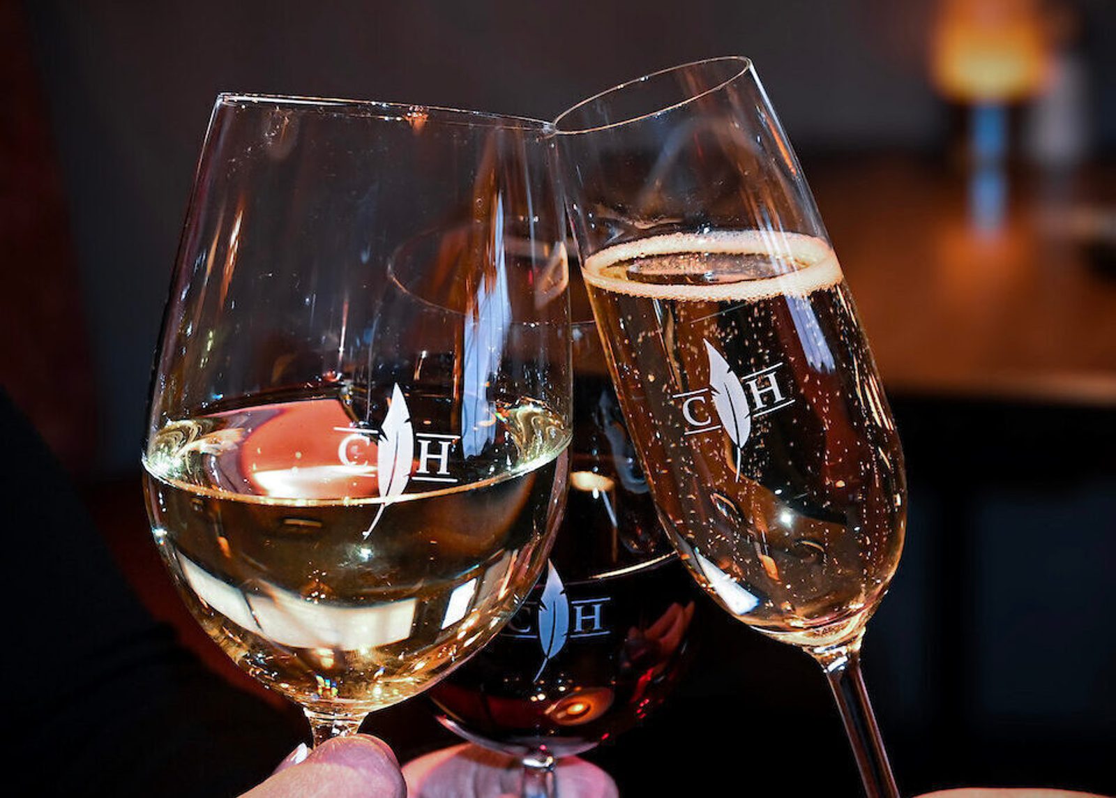 Three wine glasses clinking together, one filled with white wine, one with sparkling wine, and one with red wine, against a softly blurred bar background