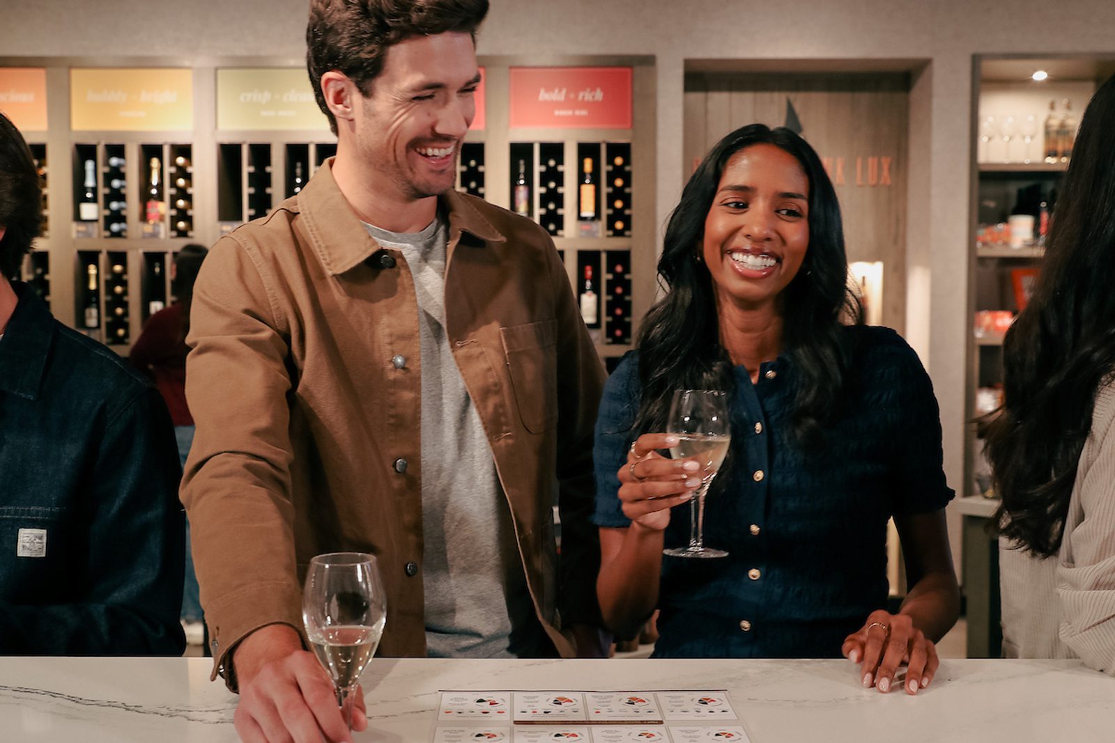 Two people smiling at a wine tasting counter, each holding a wine glass, with wine bottles displayed on shelves behind them.