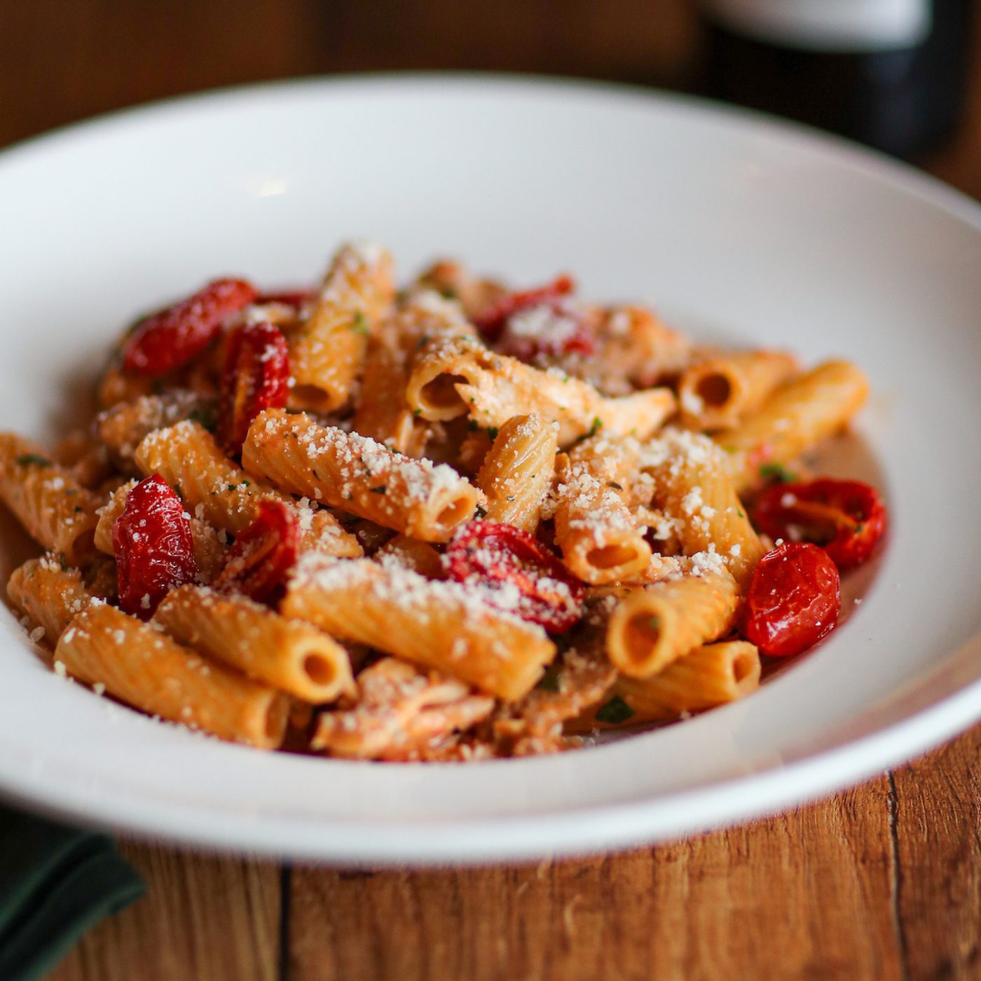 Bowl of rigatoni pasta with cherry tomatoes, herbs, and grated cheese, served in a white dish on a wooden table.