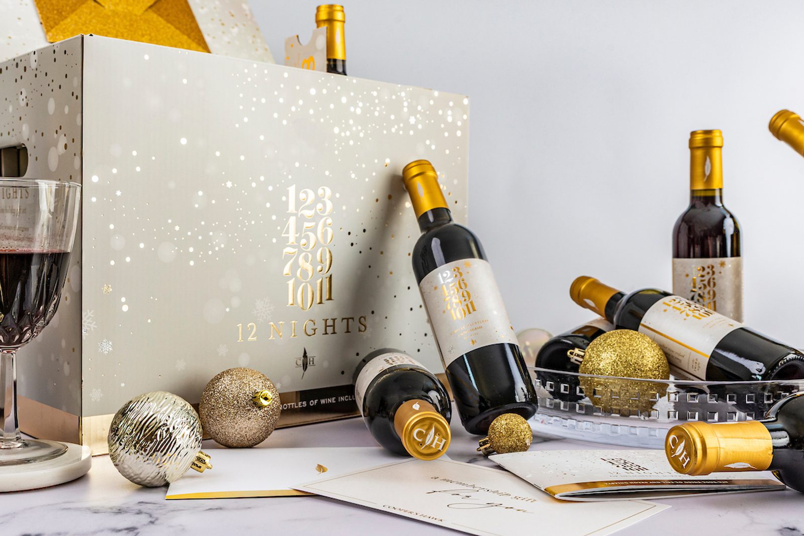 Festive wine advent calendar with several red wine bottles, a filled glass, gold ornaments, and a “12 Nights” gift box on a white tabletop