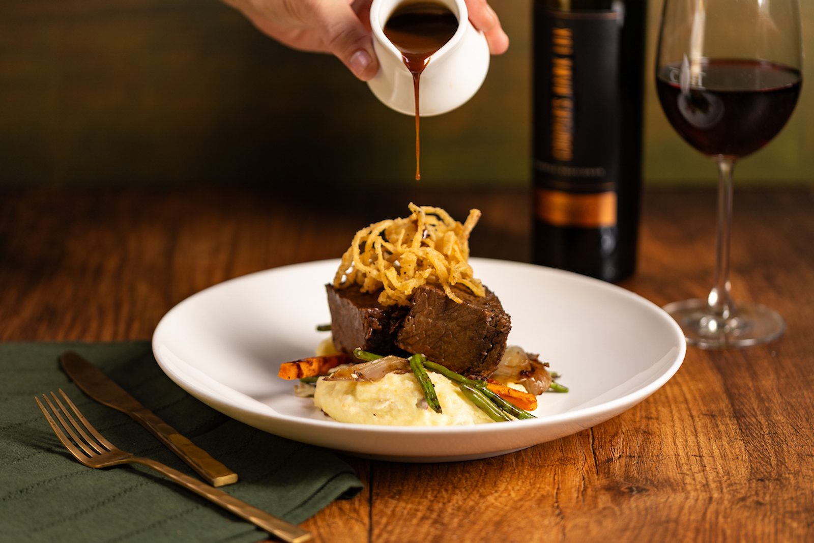 Hand pouring rich sauce over a plated beef entrée with mashed potatoes, vegetables, and fried onions, beside a glass of red wine on a wooden table