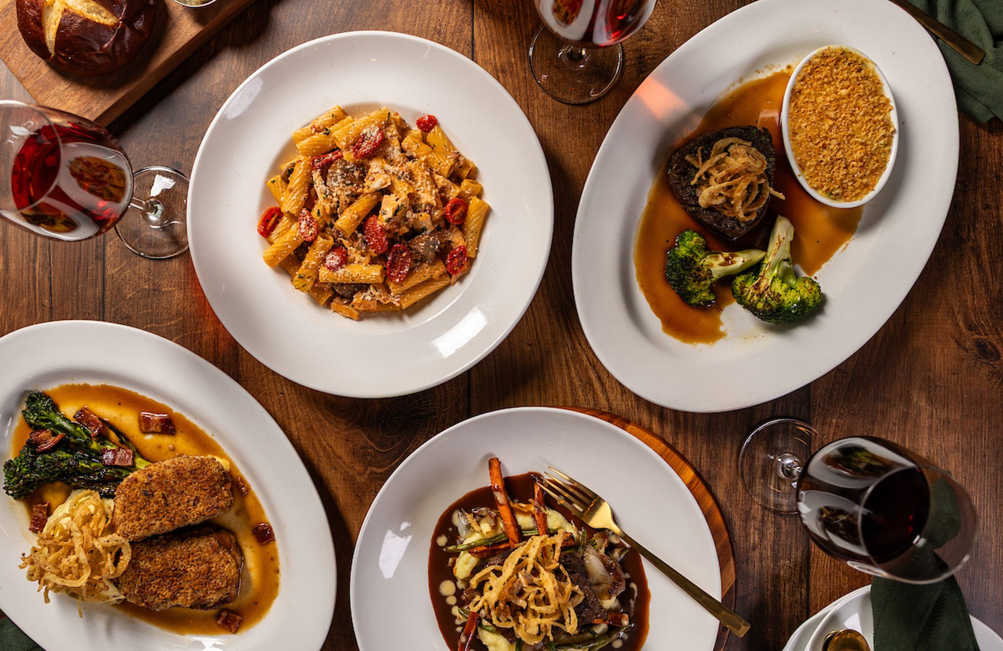 Overhead view of a festive dinner table with plated pasta and meat dishes, bread, wine glasses, green napkins, and gold cutlery on a wooden table
