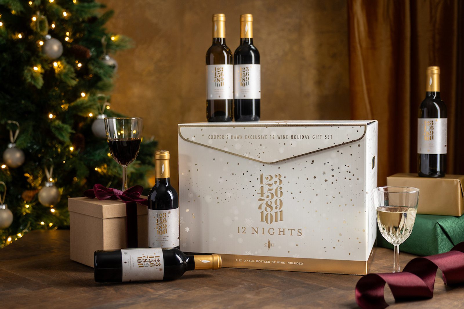 Holiday wine gift set with several bottles and wine glasses beside wrapped presents, set near a decorated Christmas tree with warm lights.