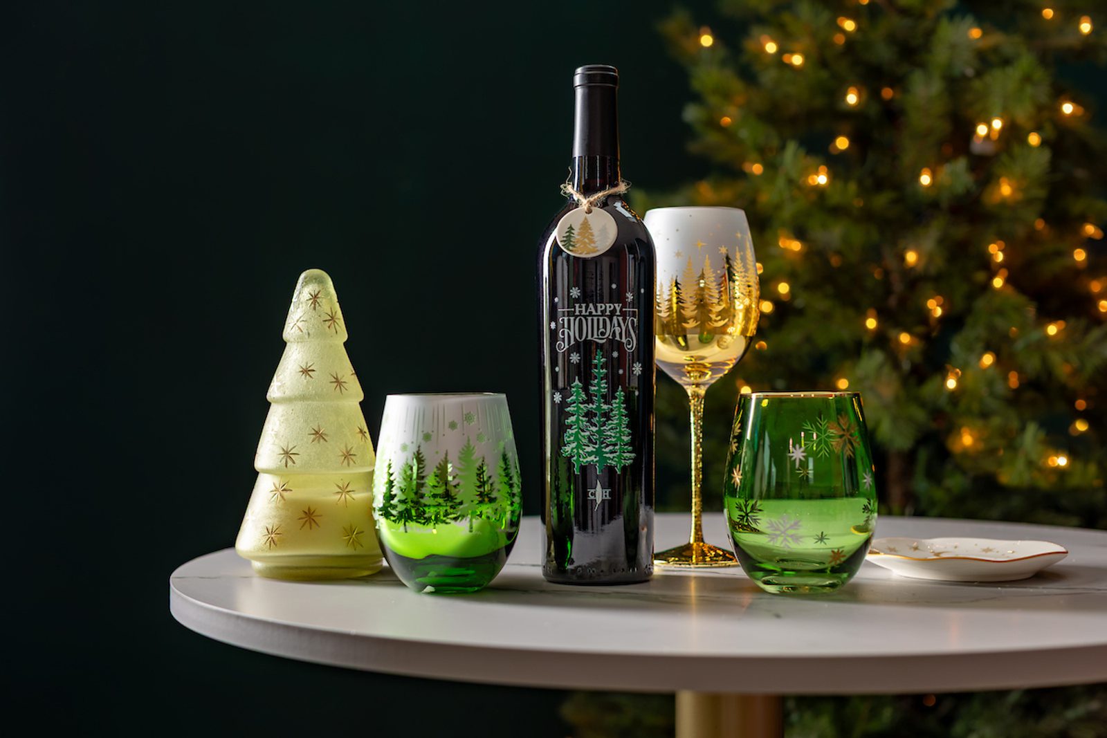 Holiday wine bottle and assorted glasses with Christmas tree designs on a round table, with a decorative tree and warm lights in the background