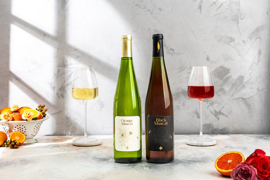 Two Muscat wine bottles labeled "Orange Muscat" and "Black Muscat" between white and red wine glasses, citrus and roses.