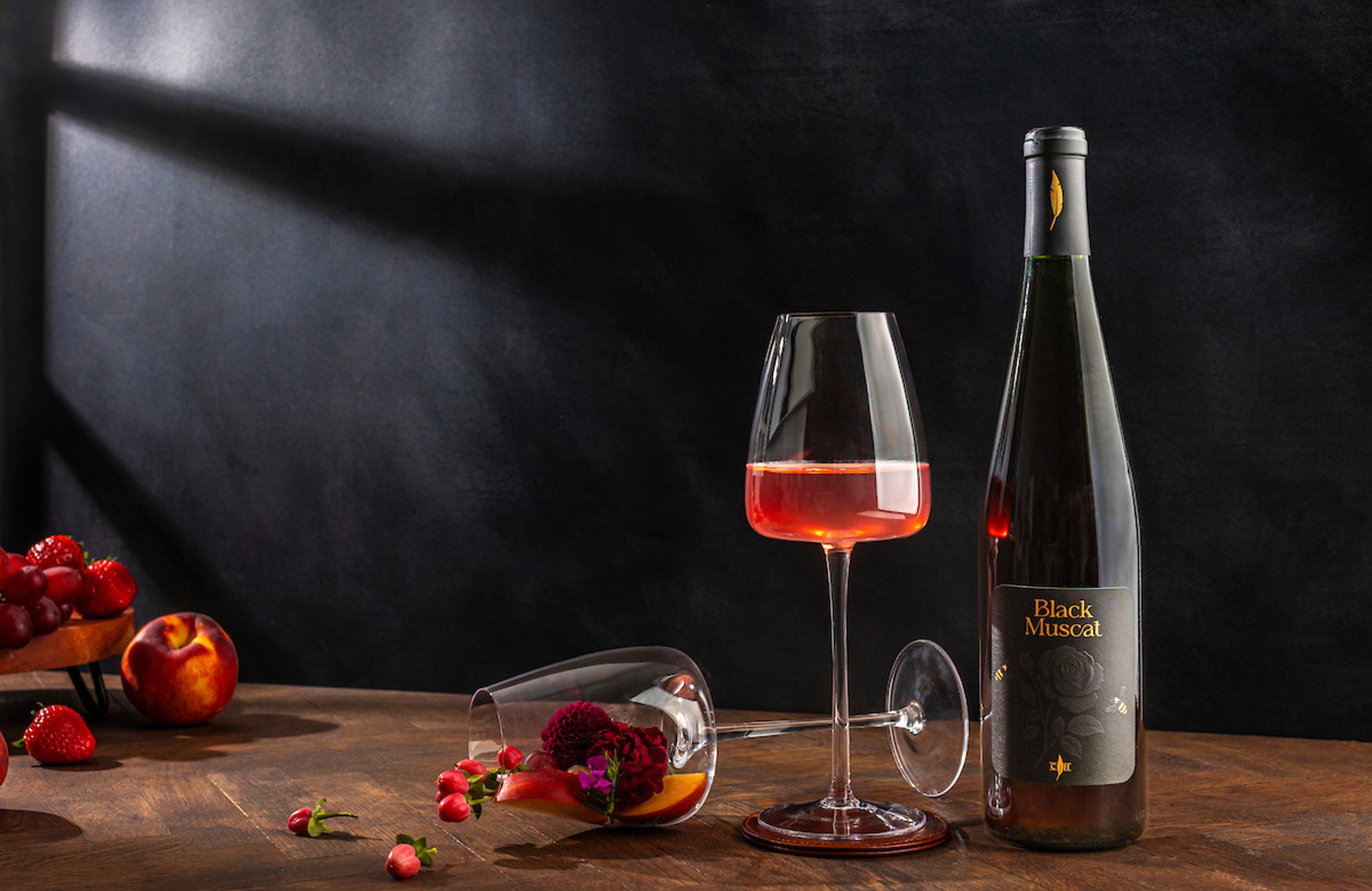 Bottle labeled Black Muscat beside a wine glass half-filled with rosé and a tipped glass with flowers and fruit on a table
