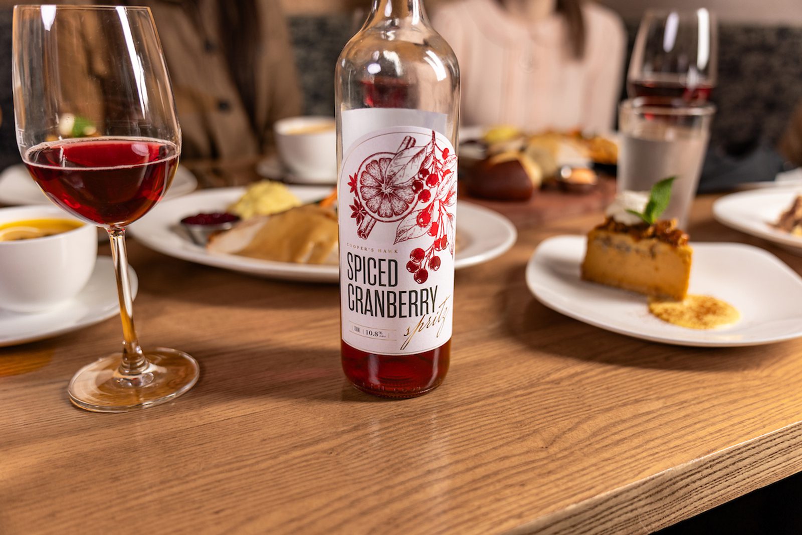 Glass of red spiced cranberry wine beside a labeled bottle on a dining table with plates of dessert and blurred people in the background