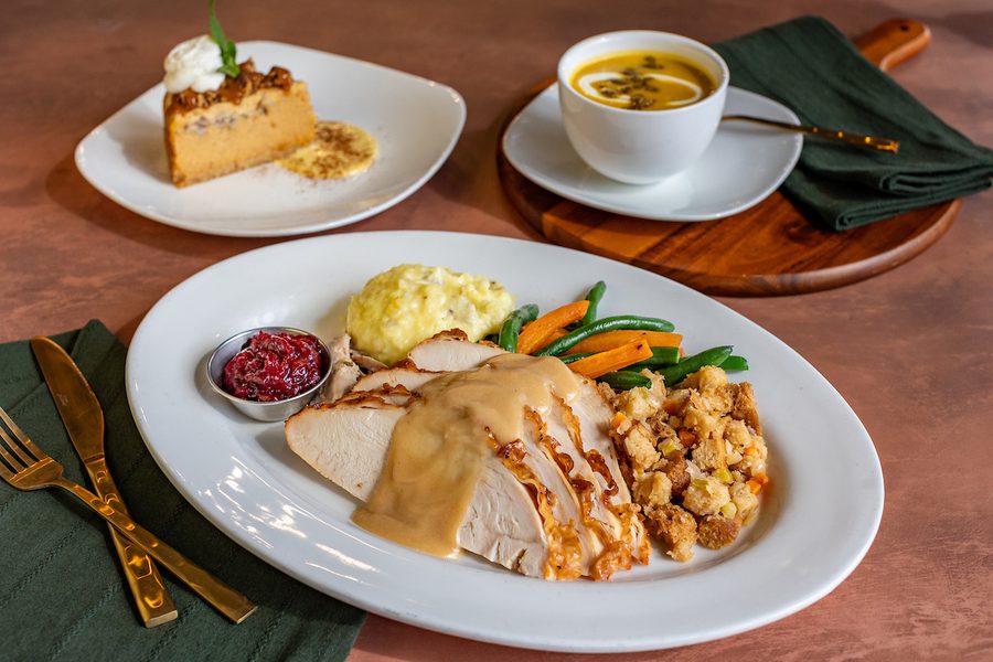 Plate of sliced turkey with gravy, mashed potatoes, green beans, carrots, stuffing, and cranberry sauce, with pumpkin soup and dessert on the side