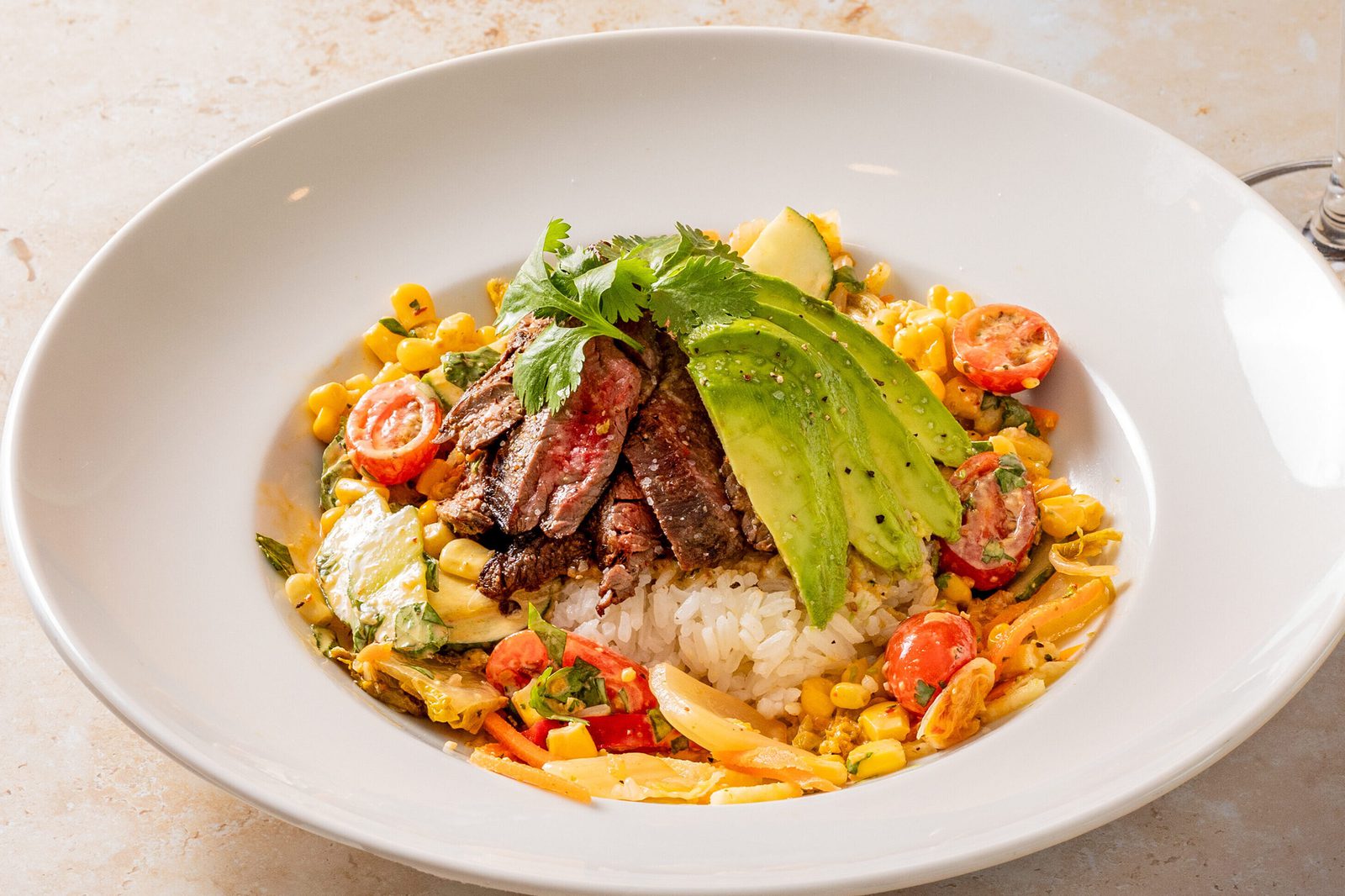 Grilled steak slices and avocado over rice with corn, tomatoes, and cilantro in a white bowl on a light stone table