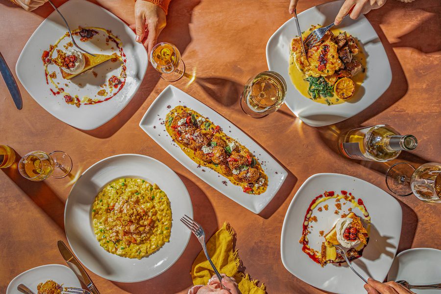 Overhead view of a restaurant table with several plated gourmet dishes, wine glasses, and hands using forks and knives during a shared meal