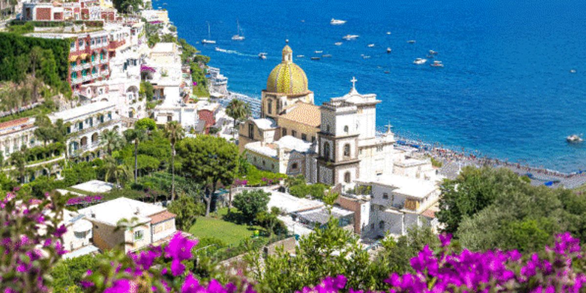 Hillside coastal town with a yellow-domed church, blue sea dotted with small boats, green trees and purple flowers in foreground