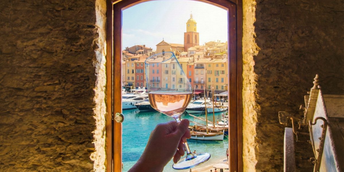 Hand holding wine glass in arched window overlooking marina with boats, beach and colorful waterfront buildings.