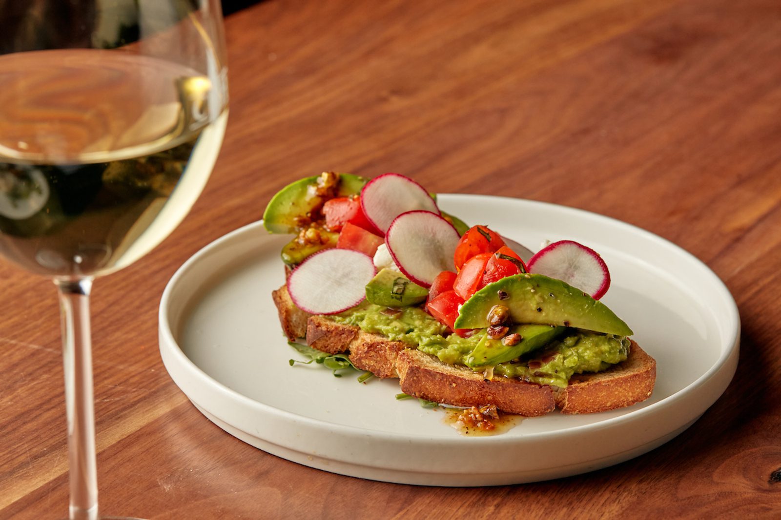 Avocado toast on a white plate topped with radish slices, cherry tomatoes, and sauce, with a glass of white wine beside it on a wooden table