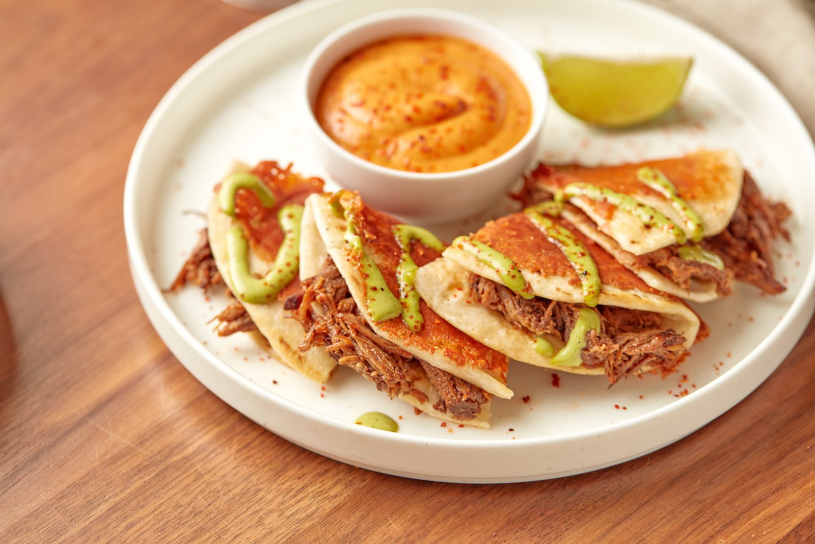 Plate of shredded beef tacos topped with green sauce, served with a cup of orange dipping sauce and a lime wedge on a wooden table