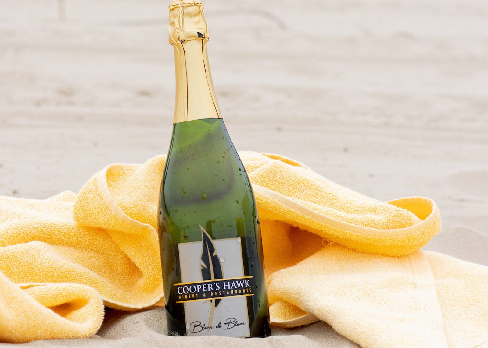 Champagne bottle on a yellow towel in sand, label reads COOPER'S HAWK WINERY & RESTAURANTS Blanc de Blanc