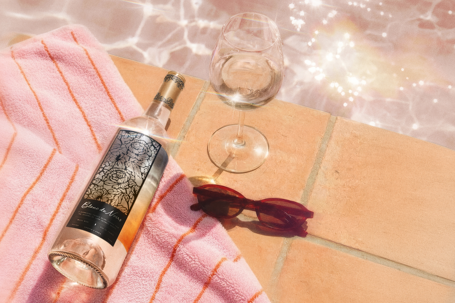Wine bottle on a pink striped towel beside a stemmed glass and red sunglasses, with sunlight shimmering across a tiled patio surface.