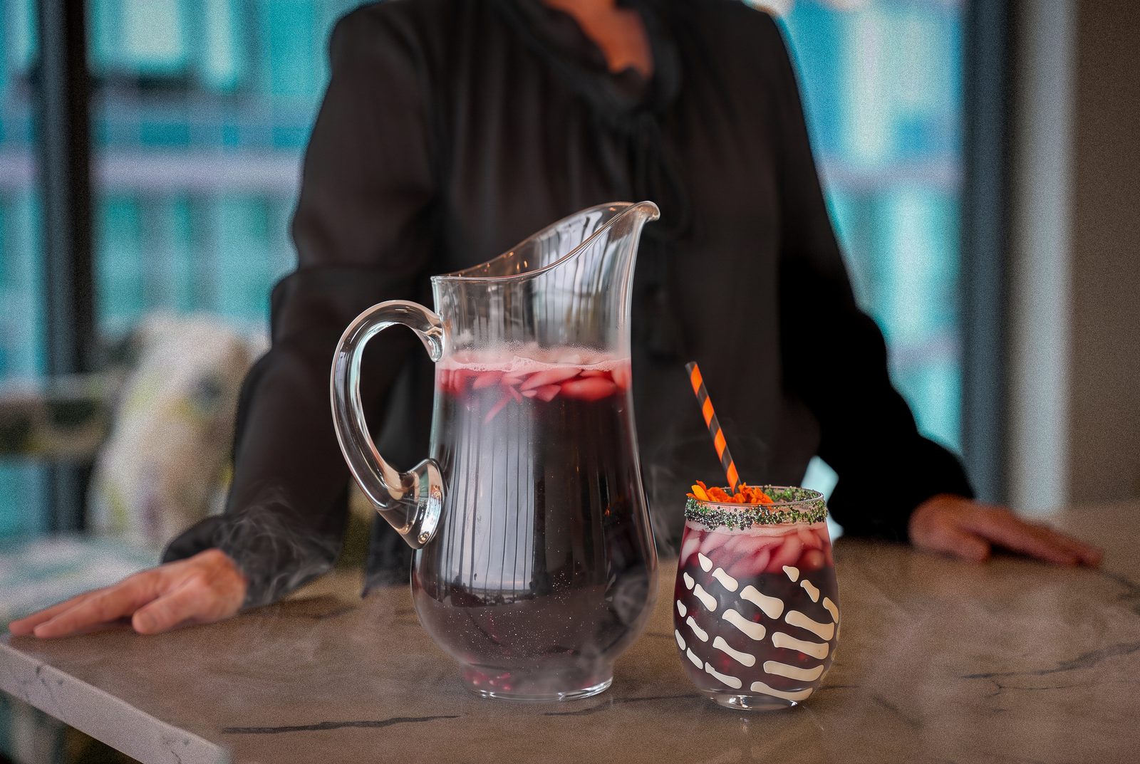 A glass pitcher of dark red drink with ice sits beside a decorated cocktail glass on a countertop, with a person in black standing behind it.