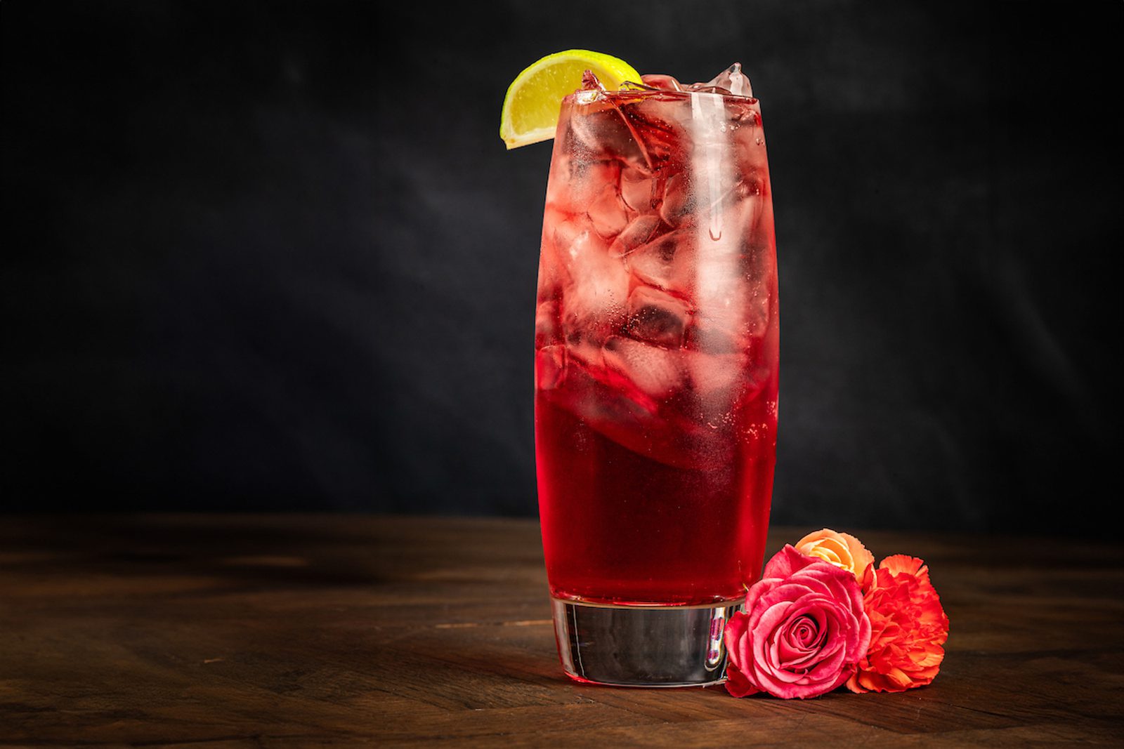 Tall glass of red iced cocktail with lime wedge on rim and pink and orange flowers on wooden table