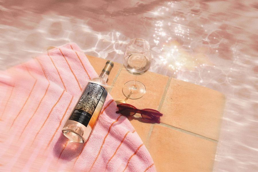 Bottle of rosé wine, a wine glass, and sunglasses on a pink striped towel beside a sunlit pool with shimmering water reflections