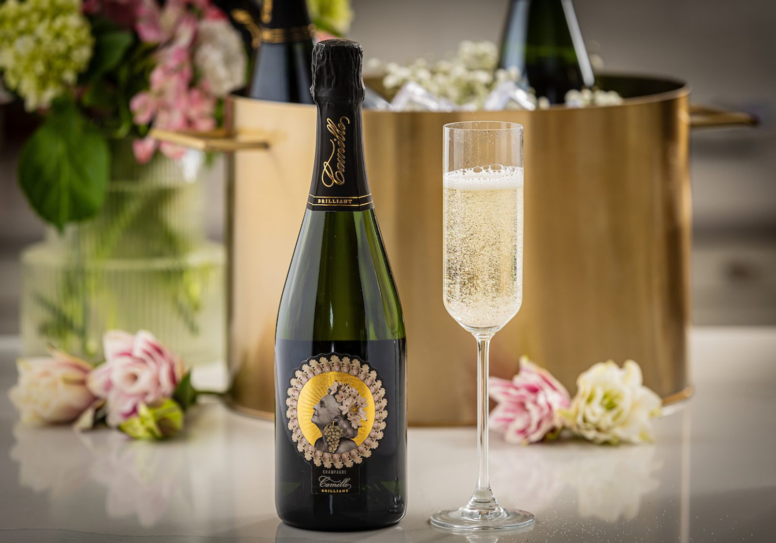 Champagne bottle with decorative floral label beside a filled champagne flute, gold ice bucket and flowers in the background