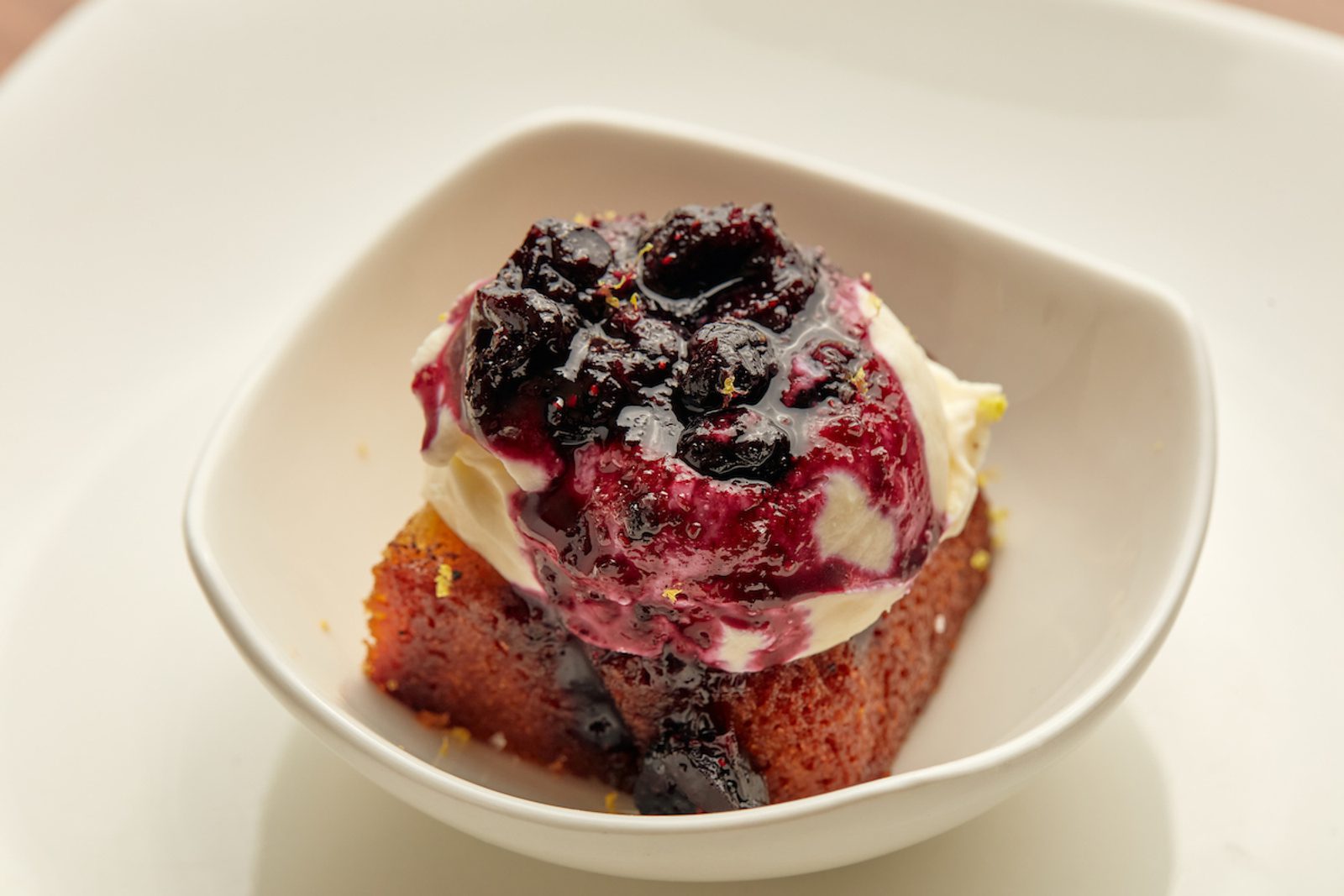 Vanilla ice cream topped with dark berry compote and a slice of cake in a white bowl, with a glossy sauce drizzled over the dessert