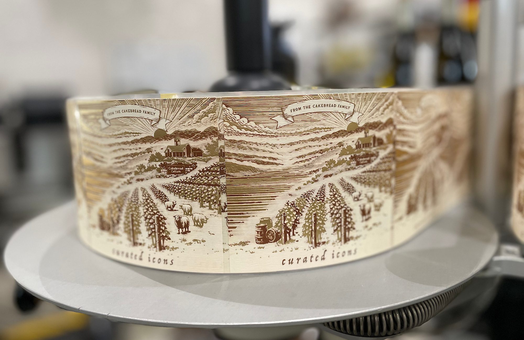 A decorated cylindrical cake with vineyard and farm illustration sits on a silver turntable, with a blurred kitchen background