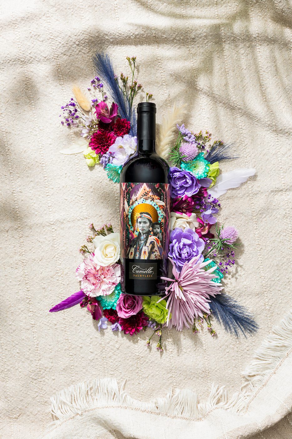 Wine bottle labeled Camille Dauntless with a colorful portrait, surrounded by mixed flowers on a beige textured blanket.
