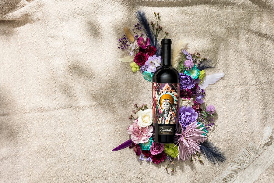 Camille Dauntless wine bottle with illustrated portrait label among purple, pink, and white flowers on beige cloth