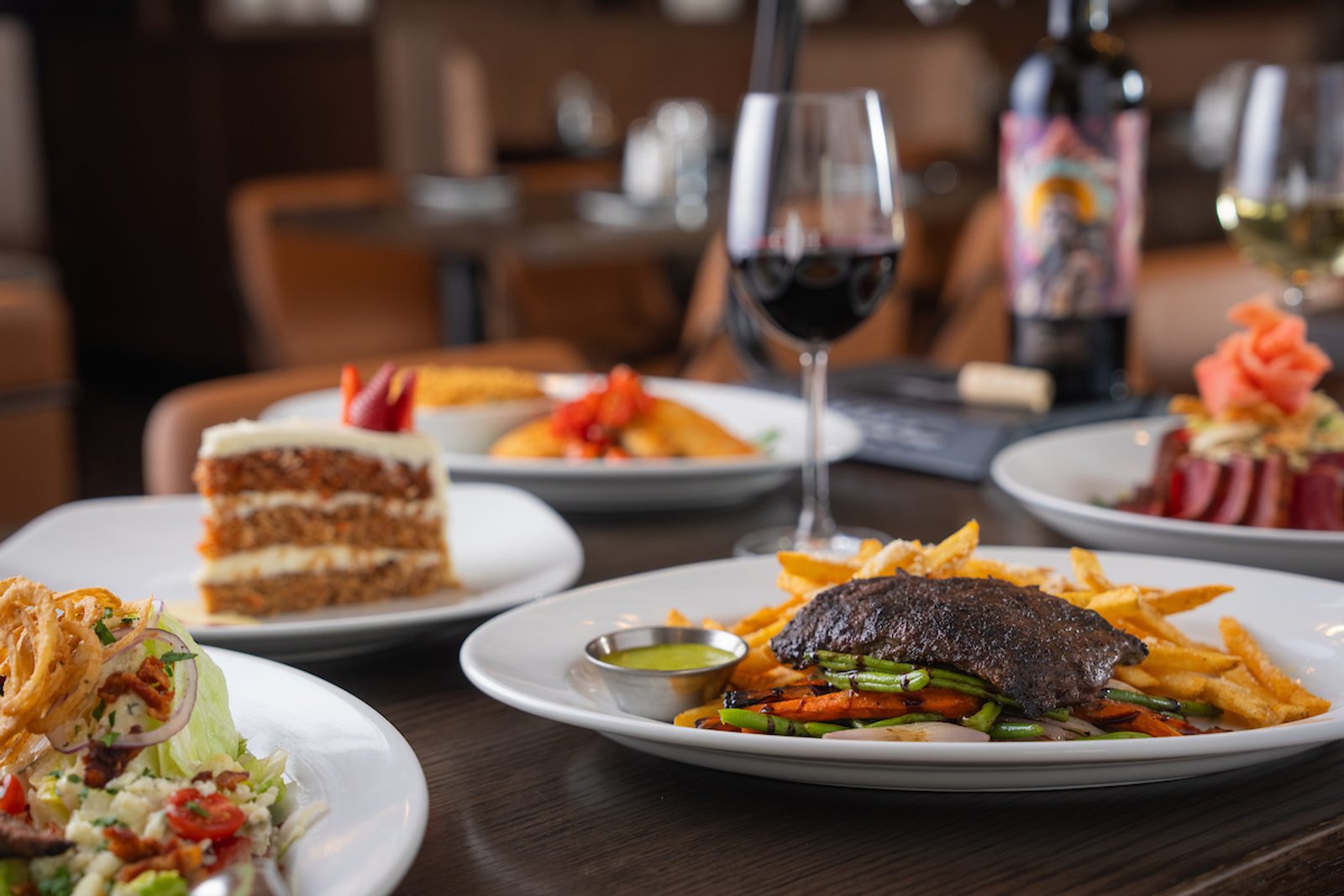 Restaurant table with assorted plated dishes, including steak and fries, salad, cake, and wine glasses in a softly blurred dining room