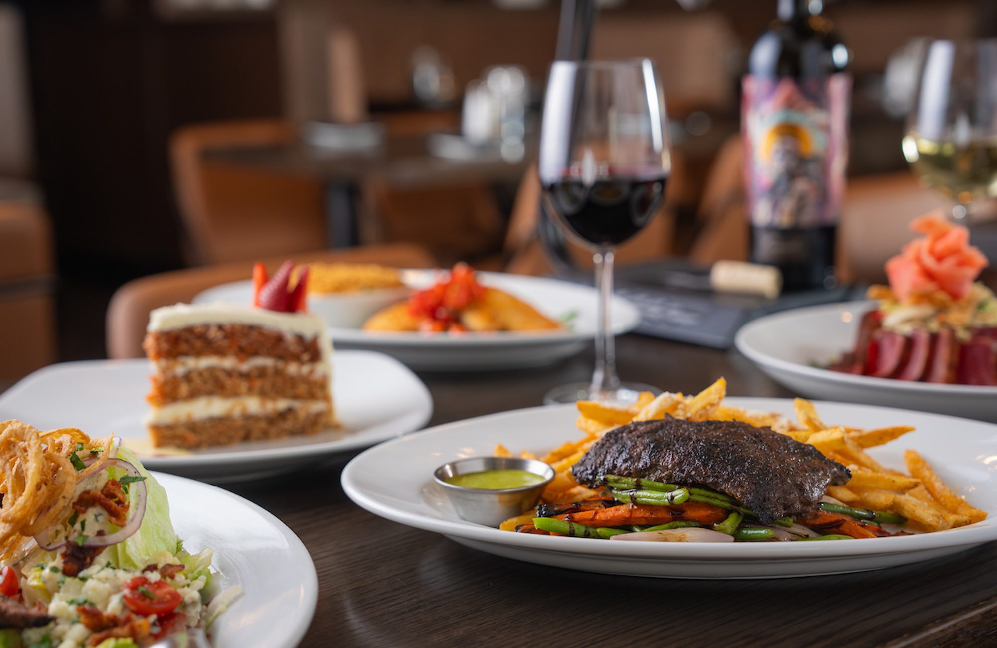 Restaurant table with assorted plated dishes, including steak and fries, salad, cake, and wine glasses in a softly blurred dining room