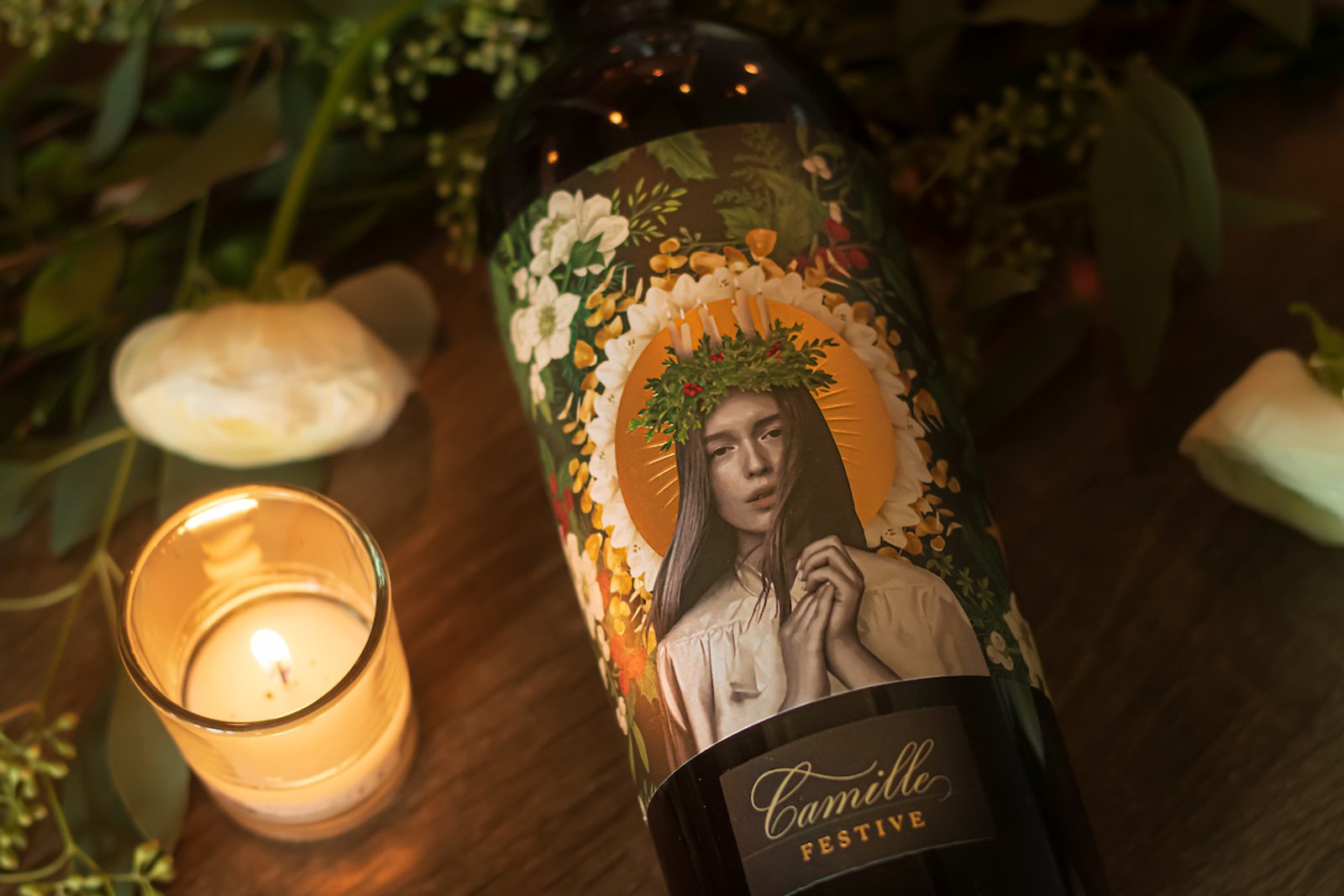 Wine bottle labeled Camille Festive with portrait of a woman wearing a wreath and floral halo, lit candle and flowers nearby