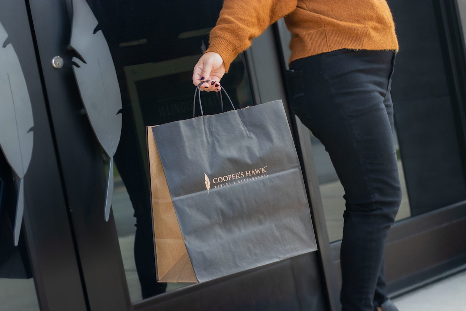 A person exiting a Cooper's Hawk restaurant is carrying a black paper gift bag with the Cooper's Hawk logo on the front.