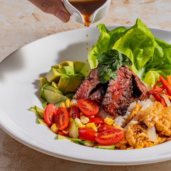 Rice bowl with sliced grilled steak, avocado, lettuce, cherry tomatoes, corn, cauliflower and sauce poured