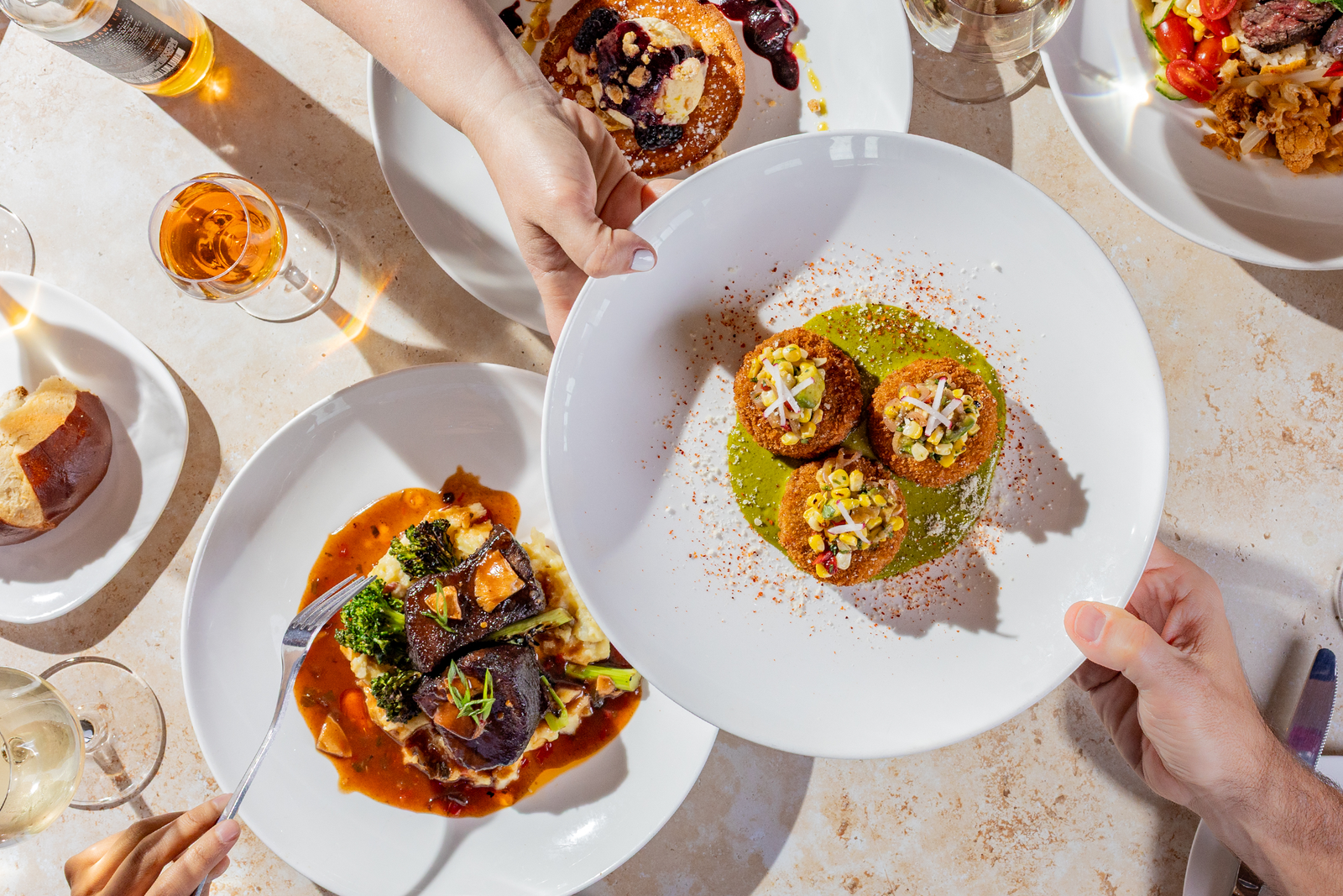 Top-down view of a restaurant table with several plated gourmet dishes, wine glasses, bread, and hands serving a white plate of three crispy appetizers over green sauce.